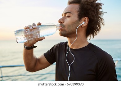 Close up portrait of Afro-American fit athlete drinking water out of plastic bottle with earphones on. Refreshing himself with water and wearing black t-shir. Taking break after hard outdoor workout