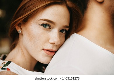 Close Up Portrait Of A Amazing Red Haired Woman With Freckles And Green Eyes Looking At Camera While Leaning Her Head On Her Boyfriend Back Outside While Dating.