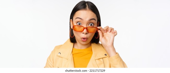 Close Up Portrait Of Asian Girl Looking Surprised, Wow Face, Takes Off Sunglasses And Staring Impressed At Camera, Standing Over White Background