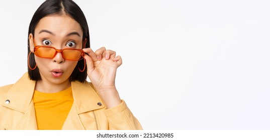 Close Up Portrait Of Asian Girl Looking Surprised, Wow Face, Takes Off Sunglasses And Staring Impressed At Camera, Standing Over White Background