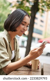 Close up portrait of asian girl sitting in cafe outside, drinking coffee and using smartphone, reading mobile phone. - Shutterstock ID 2233829793