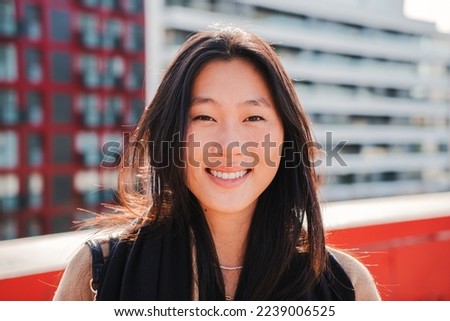 Close up portrait of asian young woman smiling and looking at camera with perfect white teeth, at the background city buildings. Attractive happy chinese female standing outdoors. High quality photo