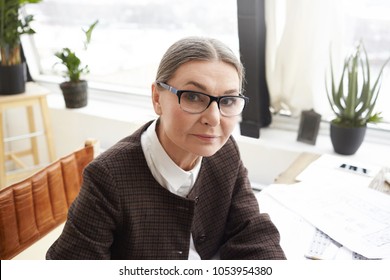 Close Up Portrait Of Attractive 60 Year Old Caucasian Woman Designer With Gray Hair Wearing Rectangular Eyeglasses Doing Paperwork In Her Light Workspace, Looking At Camera With Serious Expression