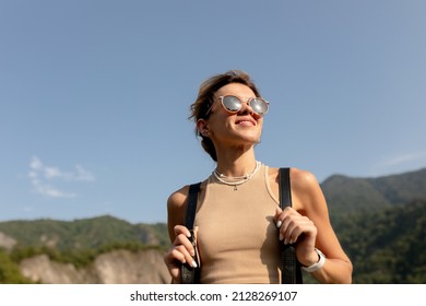 Close up portrait of attractive charming woman with collected light hair wearing sunglasses and beige t-shirt is walking aside in mountains on background of blue sky. High quality photo