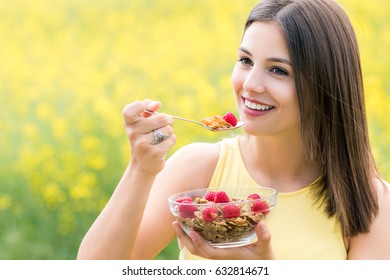 Close Up Portrait Of Attractive Healthy Young Woman Eating Crispy Whole Grain Cereal Breakfast Outdoors.Girl Holding Bowl With Cereal And Spoon Against Colorful Yellow Flower Field.