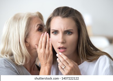 Close up portrait attractive middle aged woman whispering gossiping to ear a secret to millennial female sitting together at home. Best friends trusting relations between mother and daughter concept