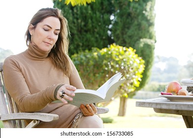 Close Up Portrait Of An Attractive Middle Aged Mature Woman Relaxing And Reading A Book In A Country Home Garden During A Sunny Autumn Morning, Near A Breakfast Table. 