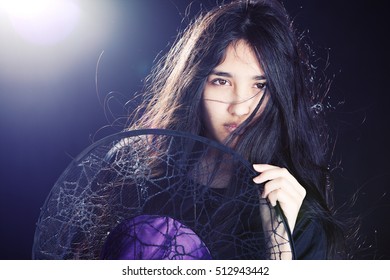 Close Up Portrait Of An Attractive Mixed Race Teen, Hair Blowing Across Her Face And Holding A Witch's Hat.  Background Light And Lens Flare Included In Image.