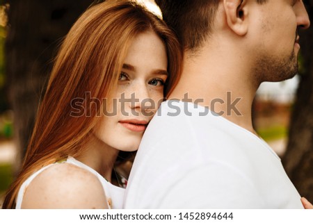 Close up portrait if an attractive red haired woman with freckles looking at camera serious while leaning her head on her boyfriends back outside.