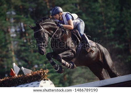 close portrait of attractive rider woman jumping over obstacle on black horse during eventing cross country competition in summer