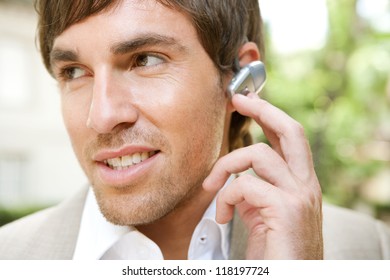 Close Up Portrait Of An Attractive Young Businessman Using A Hands Free Ear Piece Device To Make A Phone Call While In A Classic City Financial District, Outdoors.