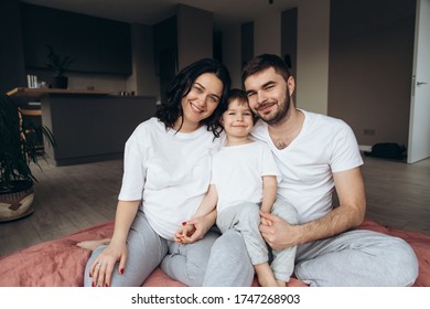 Close Up Portrait. Attractive Young Family With A Child At Home.  Smiling People. Parents And Their Blond Blue Eyes Son Are Looking To A Camera. Family Wear White T Shirts. 