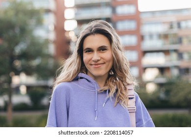 Close up portrait attractive young happy woman with fresh and clean skin stands outside city near building. Smiling curly blonde wear purple hoody and look at camera. Lifestyle, female beauty concept.