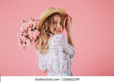 Close up portrait of an attractive young woman in summer dress and straw hat holding carnations bouquet and looking over her shoulder isolated over pink background