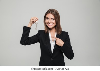 Close up portrait of an attractive young woman in suit showing key and pointing finger isolated over white background