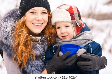 Close Up Portrait Of Baby Sitting With Young Beautiful Mother Outside At Snowy Trees Winter Background And Drinking Hot Tee. Happy Family Enjoying Beautiful Frosty Days On Christmas Picnic