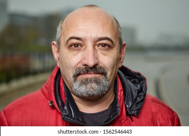 Close Up Portrait Of A Bald Man With A Gray Beard Outdoors In Cloudy Weather. Mature Adult Male In A Red Synthetic Jacket, Stands On A City Street. Businessman 50 Years Old, Tight Build, Brown Eyes.