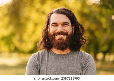 Close up portrait of bearded hipster man with long hair looking at smiling at the camera whie standing outdoor in park.