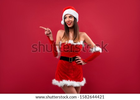 Close up portrait beautifiul caucasian woman in red Santa hat and dress on red studio background. Christmas New Year holiday concept. Cute girl teeth smiling positive Showing forefinger finger side