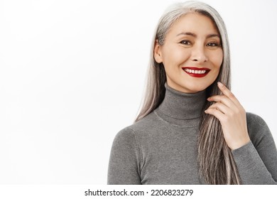 Close Up Portrait Of Beautiful Asian Female Model, Middle-aged, Touching Long Healthy Grey Hair, Smiling And Looking Happy, White Background. Aging Beauty Concept