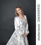 close up portrait of beautiful blonde female model wearing romantic historical gown of white silk with floral pattern and bow. Elegant standing pose, isolated on dark studio background.