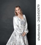 close up portrait of beautiful blonde female model wearing romantic historical gown of white silk with floral pattern and bow. Elegant standing pose, isolated on dark studio background.