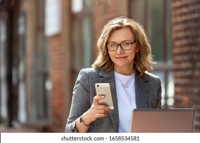 Close Up Portrait Of A Beautiful Business Woman, 55 Years Old. Use Your Cell Phone And Laptop, Outdoors, Near The Office.