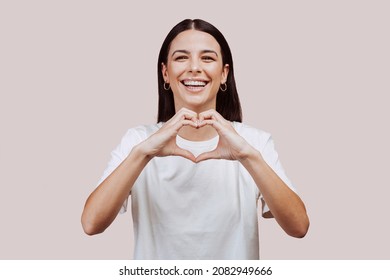 Close up portrait of beautiful caucasian young woman doing love hearth sign with hands at the camera at studio over beige background. Joyful happiness people concept.