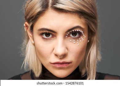 Close Up Portrait Of Beautiful Charming Young Blonde Female Model With Short Dyed Hair Staring Right At Camera, Having Mysterious Intent Gaze, Wearing Black Dress, Face Piercing And Glamorous Make Up