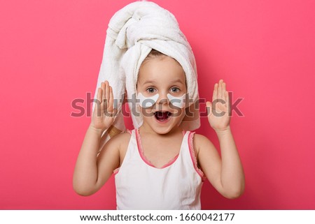 Close up portrait of beautiful excited little girl wearing white towel after shower. Studio shot isolated over pink background, female child with hands up and opened mouth, expresses astonishment.