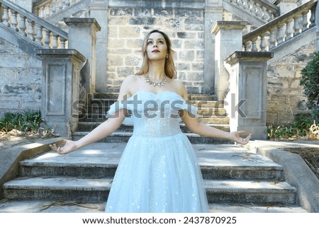 Close up  portrait of beautiful female model wearing blue fantasy ballgown, like a fairytale elf princess.  Elegant pose, gestural hands reaching out, on the terrace of a romantic castle staircase 