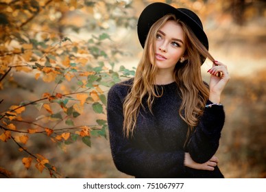 Close up portrait of a Beautiful girl in dark dress 
and black hat standing near colorful autumn leaves. Art work of romantic woman .Pretty tenderness model looking at camera.