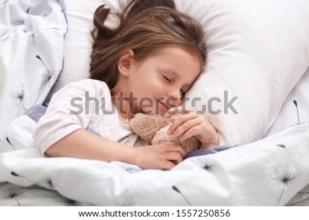 Close up portrait of beautiful girl sleeping in pajama in bed with her teddy bear, lying on pillow with closed eyes, charming cute female kid having dark hair. Childhood and morning time concept.