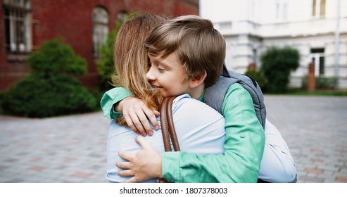 Close Up Portrait Of Beautiful Happy Caucasian Woman Hugging Her Son At Schoolyard Outdoors. Cheerful Teen Male Junior Student With School Bag Hugs Mother Near School. Love Concept