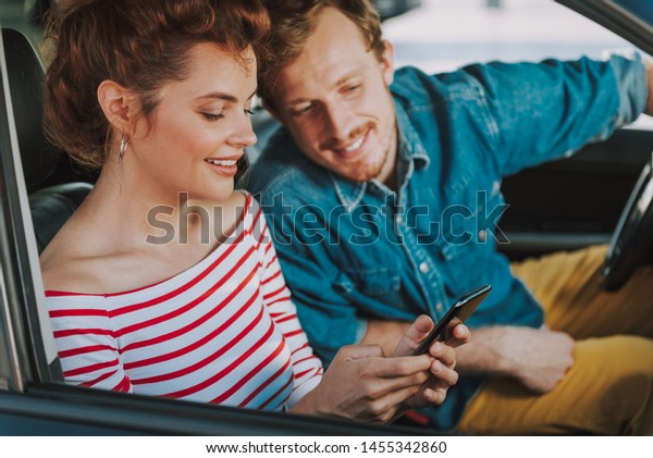 Close up portrait of\
beautiful lady using smartphone in auto. Bearded man looking at\
phone camera and smiling