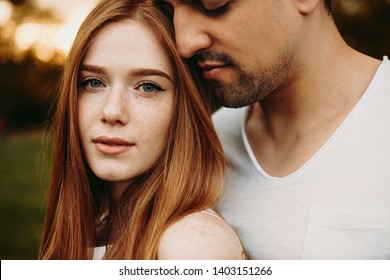 Close Up Portrait Of A Beautiful Red Hair Woman Looking At Camera While Her Boyfriend Embracing Her With Closed Eyes Near Her Face Against Sunset While Dating.