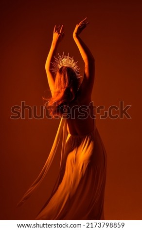 Close up portrait of beautiful red-haired woman wearing long flowing fantasy toga gown with golden halo crown jewellery,  creative hand gestures on a dark moody background with glowing orange lighting