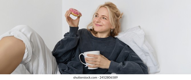 Close up portrait of beautiful smiling, blond woman, lying in bed, drinks tea and eats doughnut on a lazy weekend or day-off. - Powered by Shutterstock