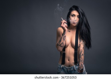 Close up portrait of beautiful woman with tattoo smoking, wearing short jeans paint against dark background.
