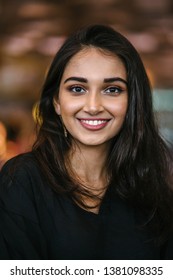 A close up portrait of a beautiful, young, attractive and photogenic Indian Asian girl during the day in the city. She is wearing a casual black dress and smiling radiantly. 