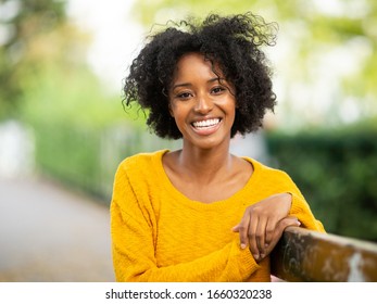 Close Up Portrait Of Beautiful Young Black Woman Sitting On Bench Outside And Smiling