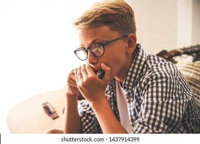 Close up portrait of beautiful young boy playing mouth harmonica. Music student exercises alone at home. Trendy caucasian child plays instruments sitting on the sofa. Blonde guy doing musical homework - Powered by Shutterstock