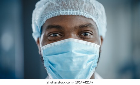 Close Up Portrait Of A Black African American Handsome Male Doctor Or Surgeon Wearing A Protective Face Mask And Disposable Surgical Cap. Scientist Calmly Looking At Camera. Patient Before Surgery.