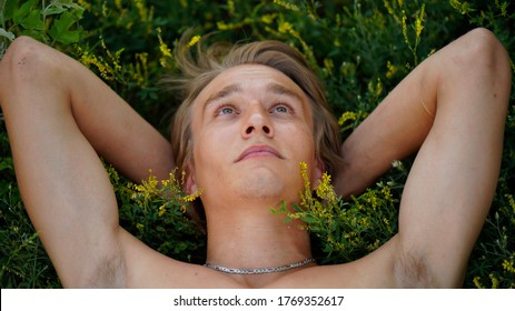 Close up portrait of blond smiling young man with chain lying in grass in park. Guy folded hands behind head