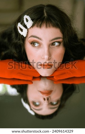 Close up portrait of a brunette caucasian woman, making faces and different expressions with her face reflected on a mirror. Pale white skin and orange vibrant gloves. Creative, artistic make up. 