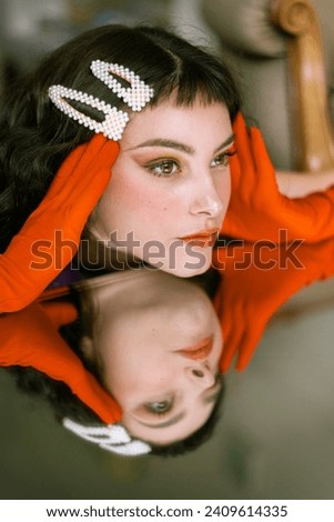 Close up portrait of a brunette caucasian woman, making faces and different expressions with her face reflected on a mirror. Pale white skin and orange vibrant gloves. Creative, artistic make up. 