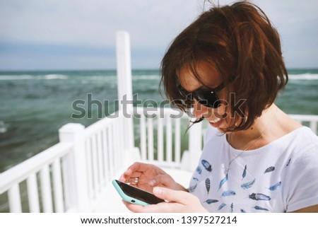 Close up portrait of brunette woman, adult woman in black sunglasses looking to mobile phone and smiling on black sea background 