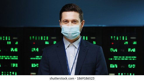 Close up portrait of Caucasian handsome male professional security engineer in medical mask stan ding in datacenter and looking at camera. Young happy man working in data storage. Server room concept