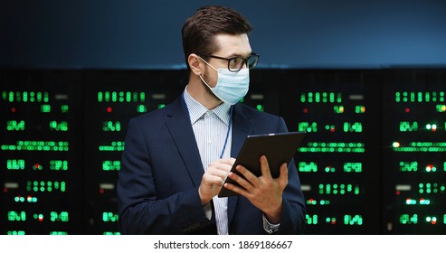 Close up portrait of Caucasian man wearing mask standing among servers with tablet device, tapping on screen and checking big data processors. Male analytic in data storage working on digital security