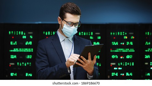 Close up portrait of Caucasian man wearing mask standing among servers with tablet device, tapping on screen and checking big data processors. Male analytic in data storage working on digital security